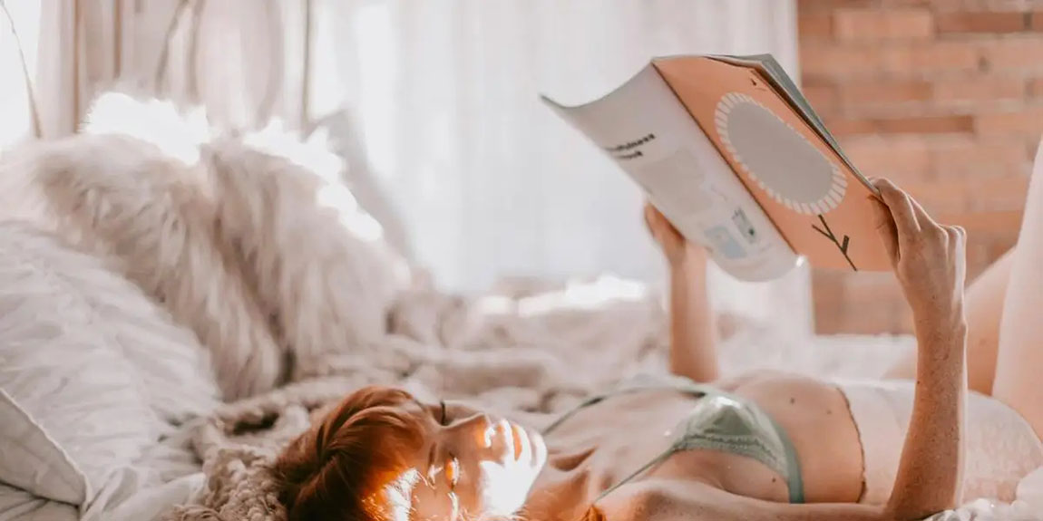 A-woman-in-lingerie-reading-a-magazine-while-lounging-on-a-bed-enveloped-in-soft-natural-light-by-Leah-Newhouse.webp.jpg