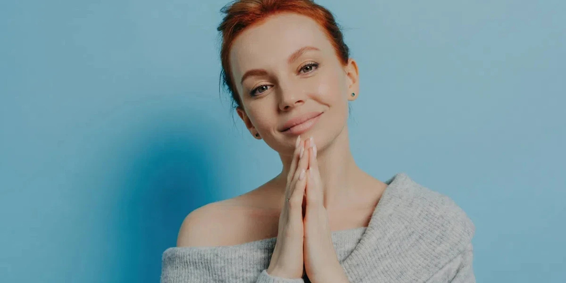 Serene-redhead-woman-with-hands-together-off-shoulder-sweater-against-a-blue-background.webp.jpg