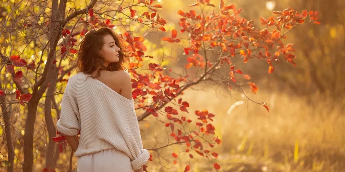 A-young-brunette-woman-in-a-cozy-off-shoulder-sweater-poses-near-a-tree-with-vibrant-autumn-leaves.webp.jpg