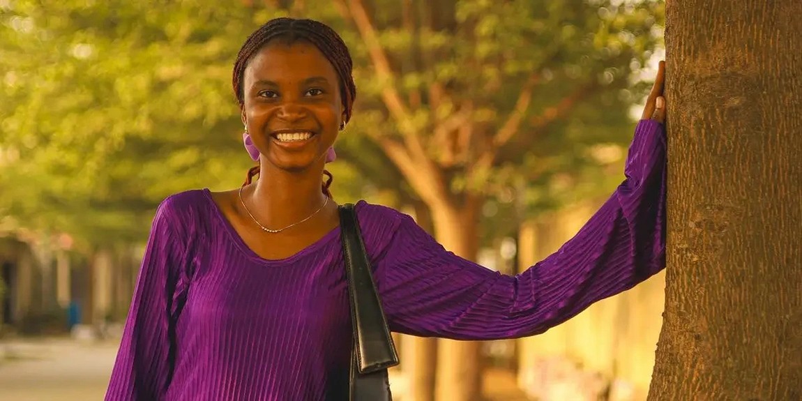 Smiling-woman-wearing-a-purple-dress-leans-on-a-tree-in-a-serene-outdoor-setting-by-Paul-Uchechukwu.webp.jpg